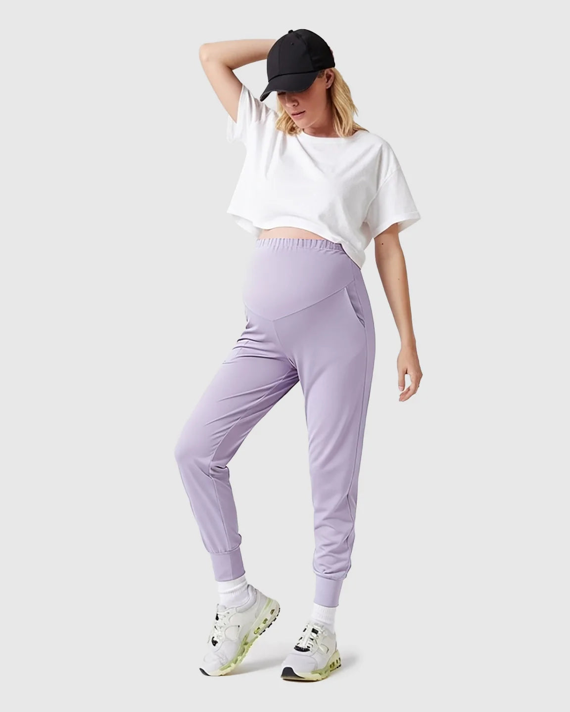 Woman in supportive slim-fitting lavender maternity joggers with a high-waist design, white crop top, and black cap.