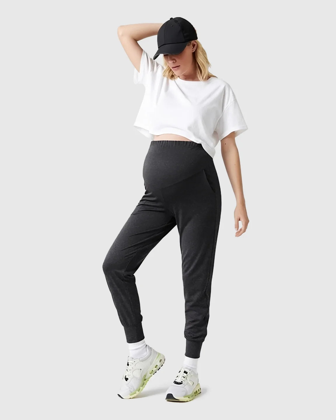 Woman in black supportive slim-fitting maternity joggers with high-waist design, white crop top, and black cap.