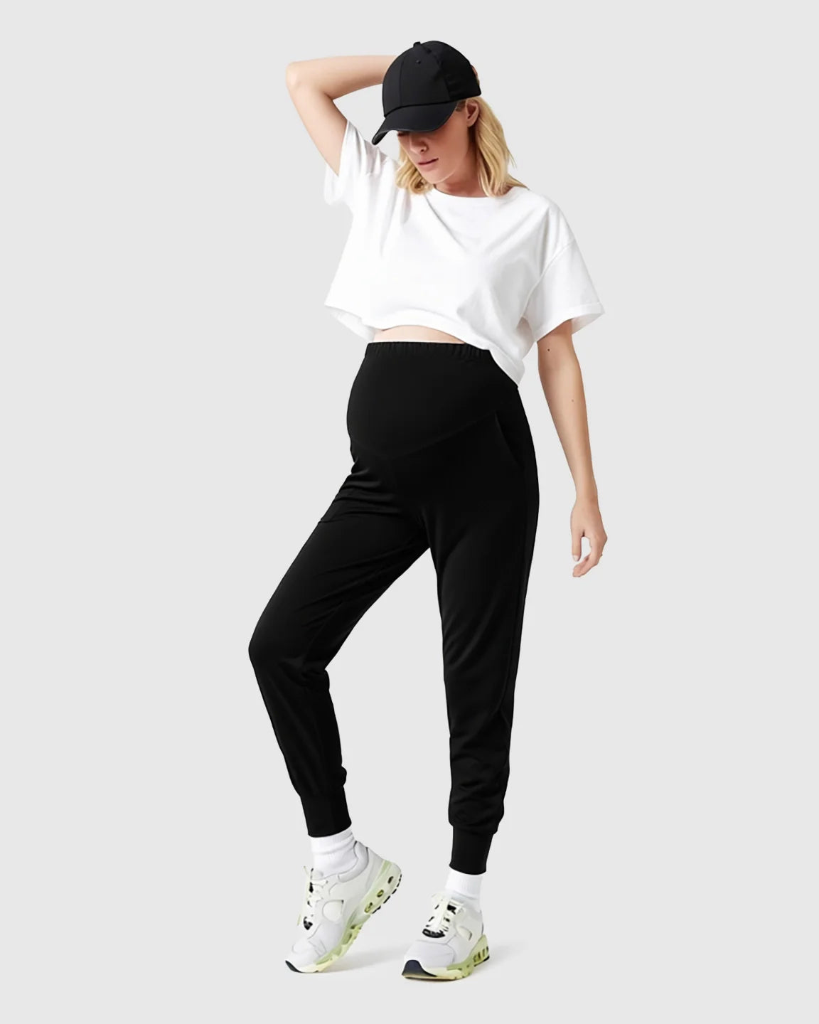 Woman in black supportive slim-fitting maternity joggers with a high-waist design, white crop top, and black cap.