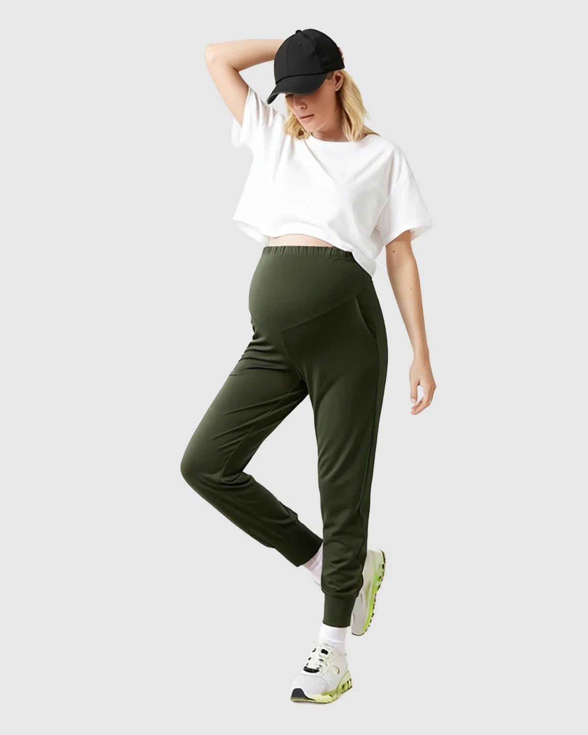 Pregnant woman in olive green supportive slim-fitting maternity joggers, white crop top, and black cap.