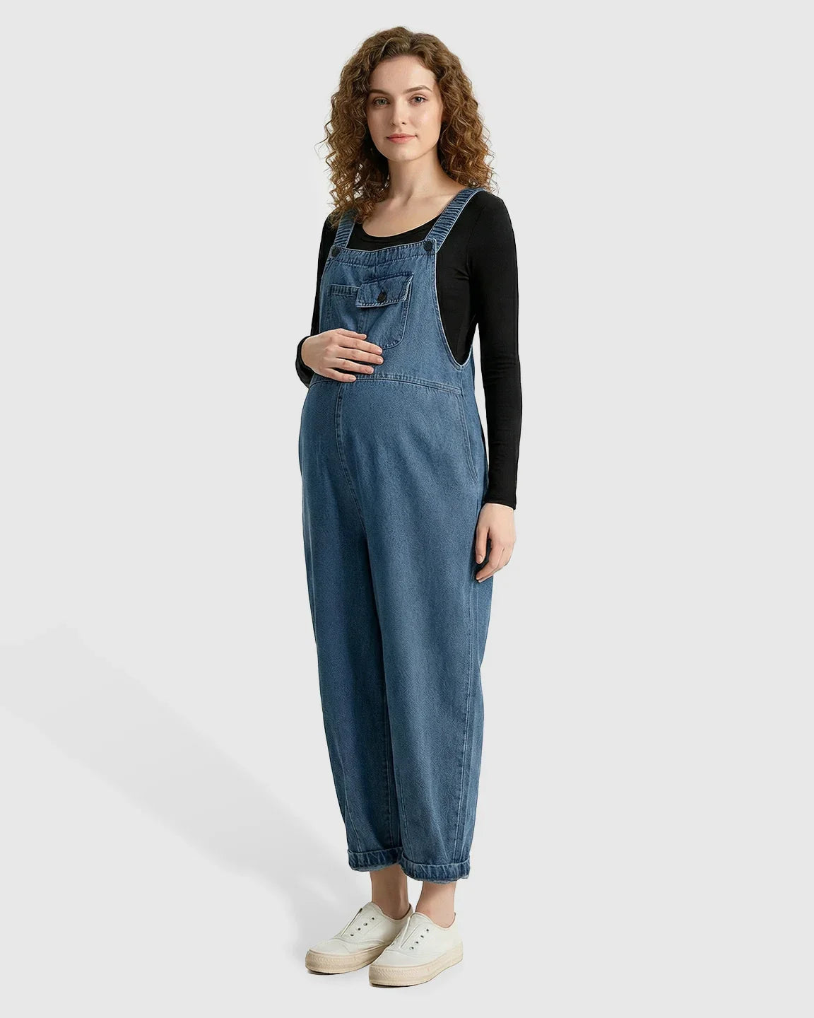 Pregnant woman in loose-fit blue denim maternity overalls with adjustable straps and rolled cuffs.