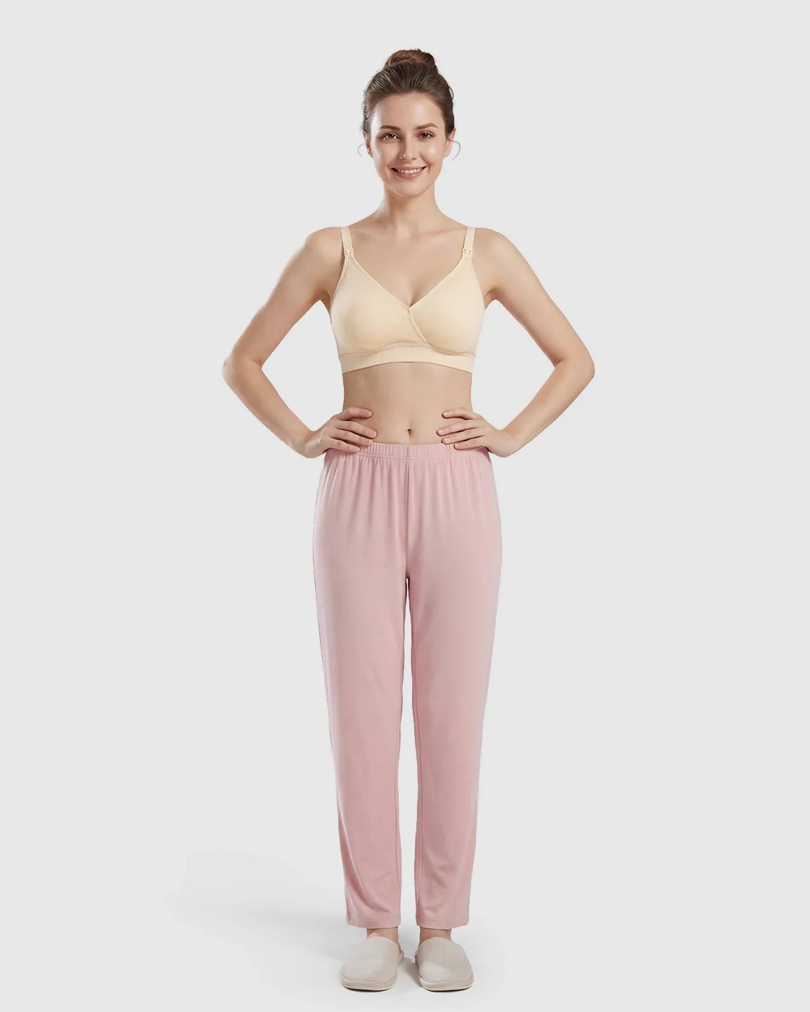 Woman models a seamless, wireless, cross-front beige nursing bra with pink lounge pants and slippers.