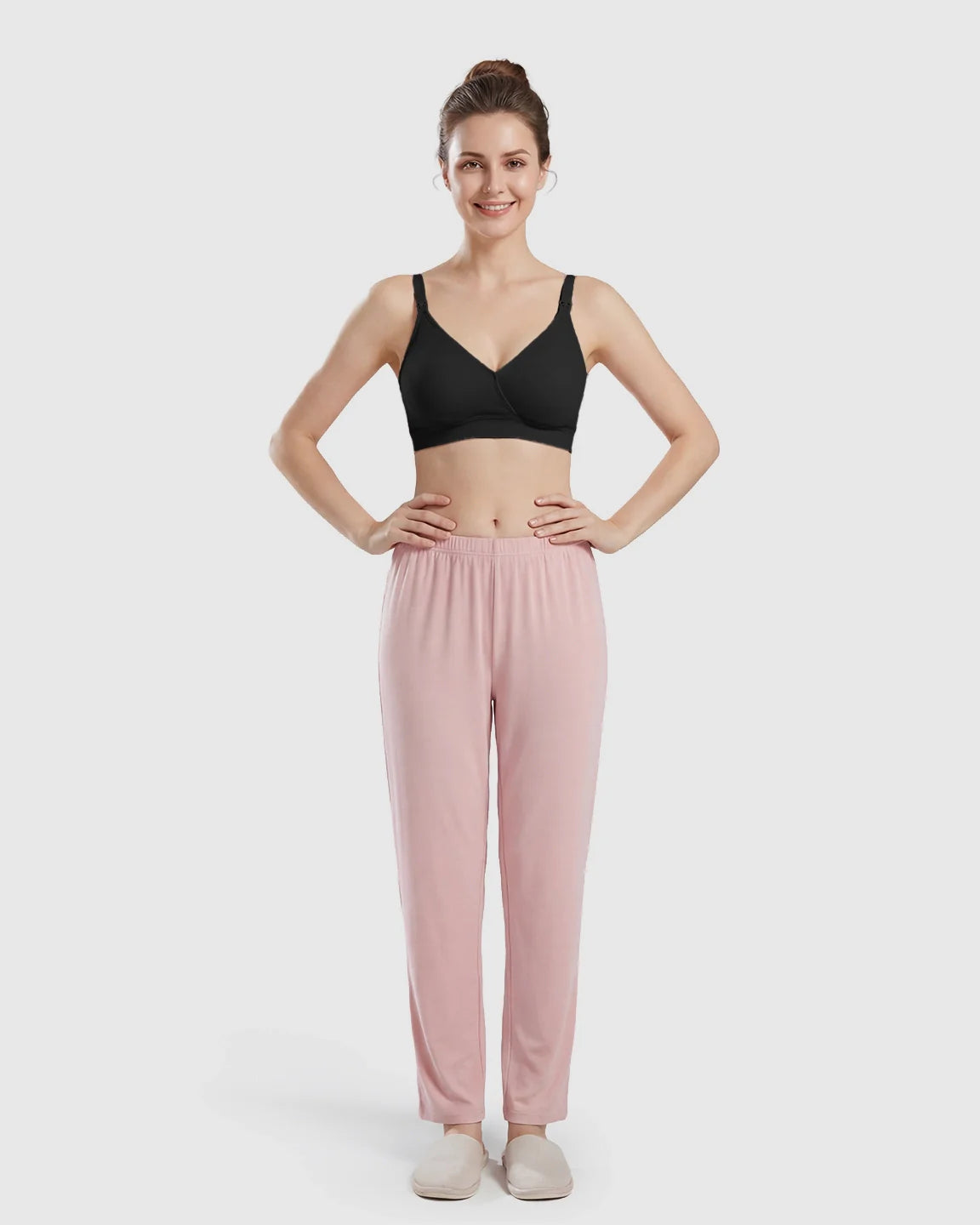 Smiling woman models a black seamless cross-front wireless nursing bra with pink pants.