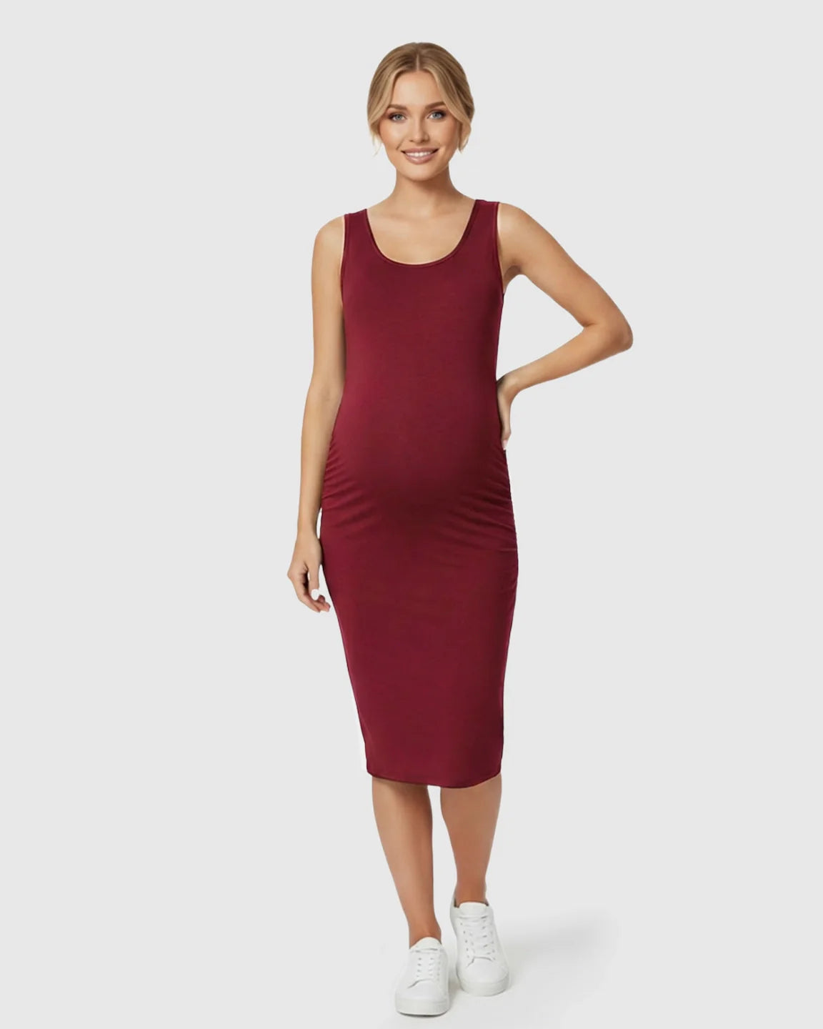 Smiling pregnant woman in a sleeveless, burgundy bodycon maternity dress with side pleats and white sneakers.