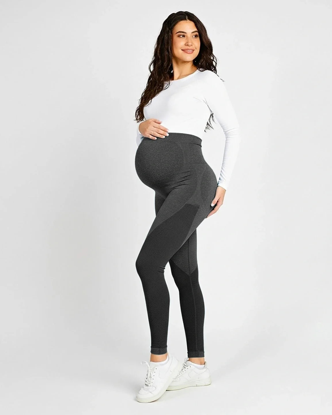Smiling pregnant woman in charcoal high-waisted maternity leggings with mesh panels and a white long-sleeve top.