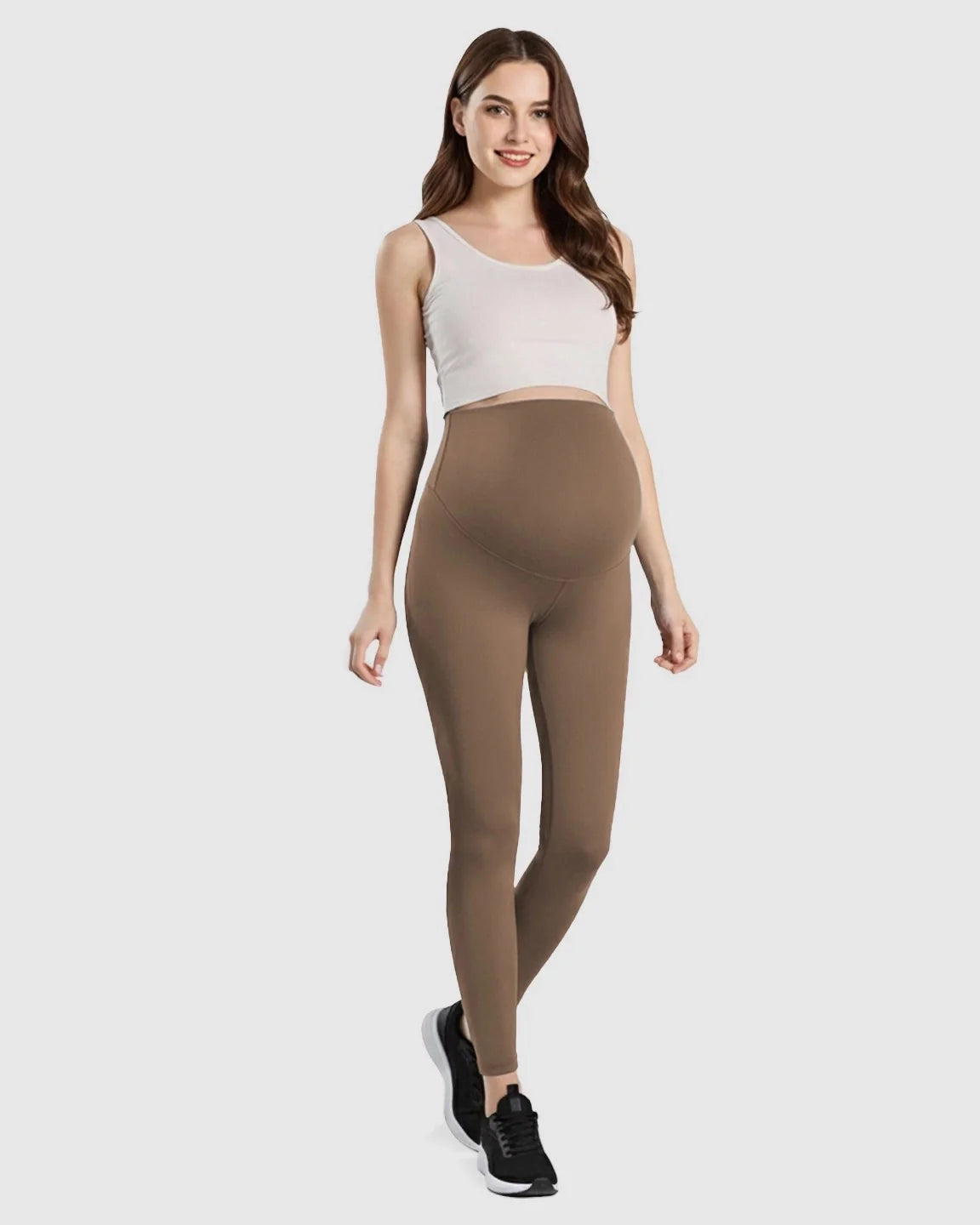 Smiling pregnant woman in brown double-layer belly support maternity leggings and a white crop top.