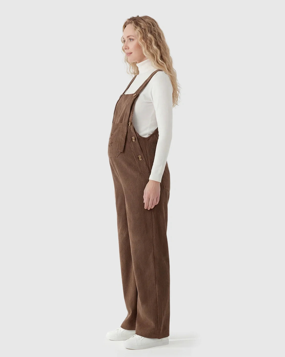 Pregnant woman in brown corduroy maternity overalls with adjustable straps over a white turtleneck.