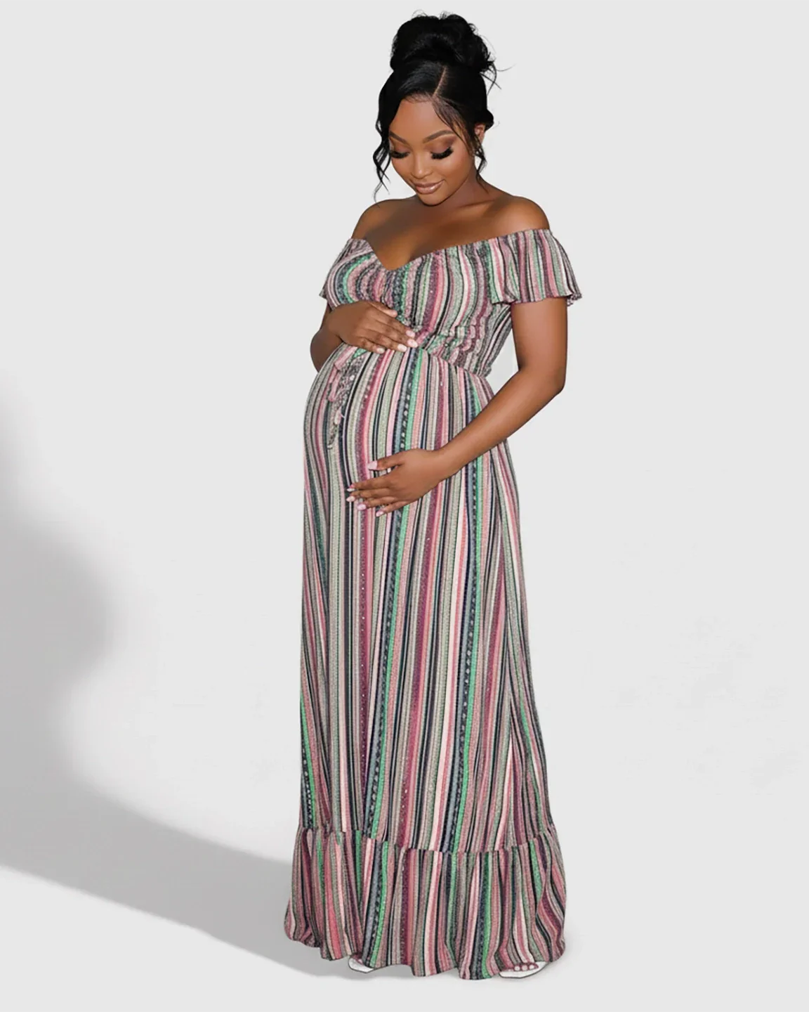 Pregnant woman in a colorful striped off-shoulder floor-length maternity maxi dress, hands on bump.