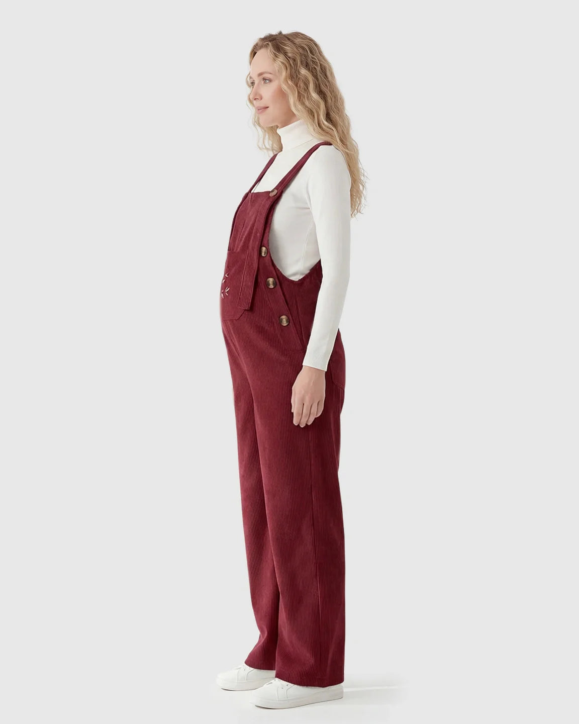 Pregnant woman in side profile wearing red corduroy maternity overalls with adjustable straps over a white turtleneck.