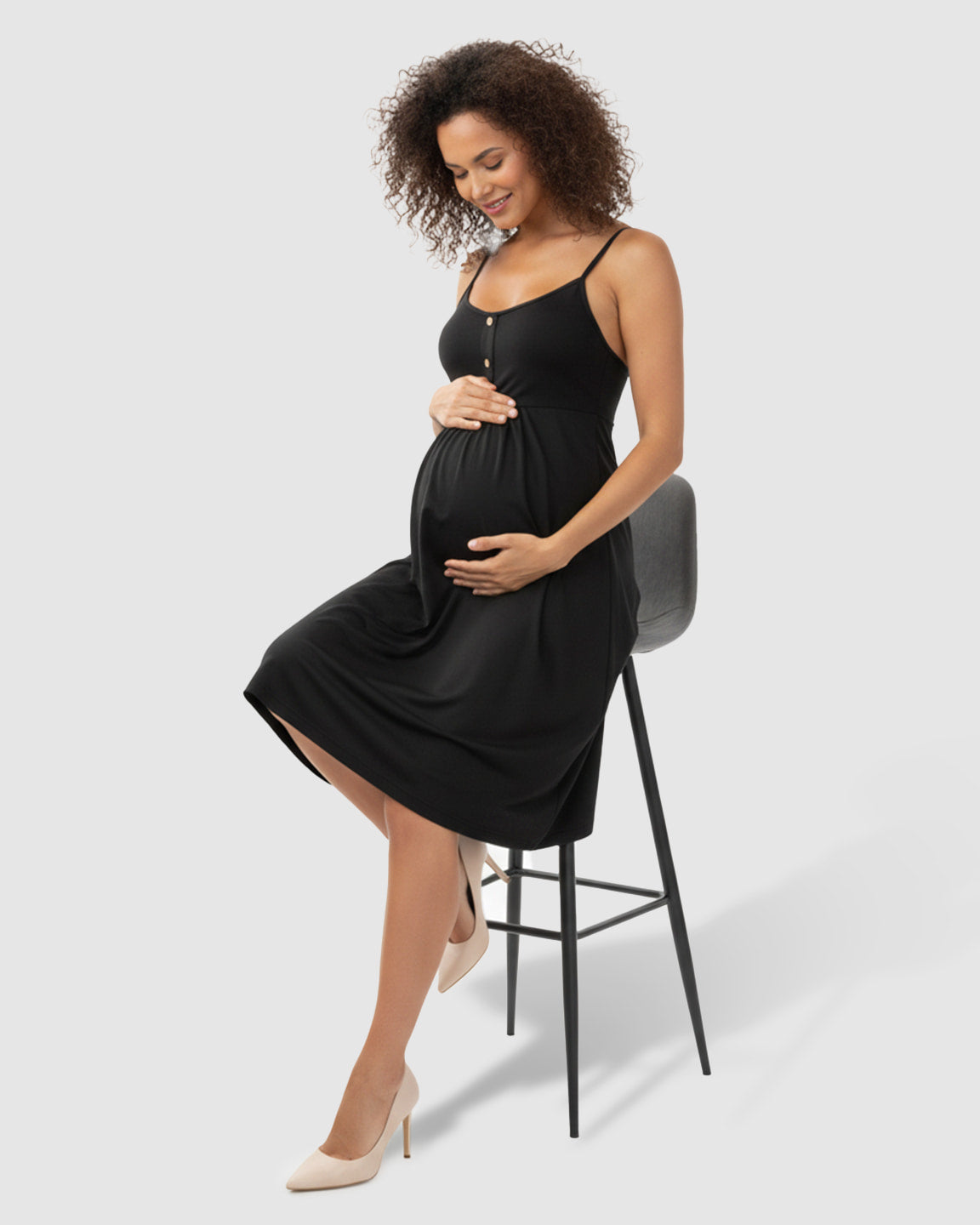 Pregnant woman in a black thin-strap A-line maternity midi dress with buttons, holding her belly.