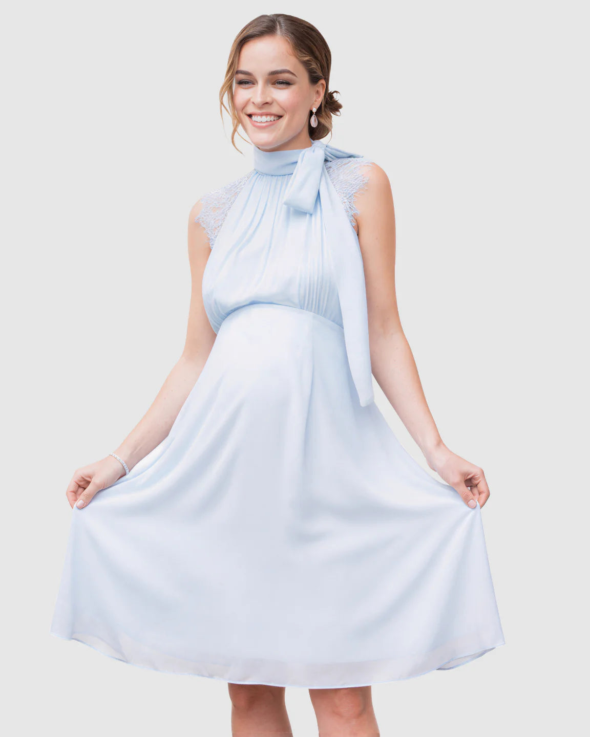 Smiling pregnant woman in a light blue chiffon high-neck midi dress with lace detail and a large tie bow.