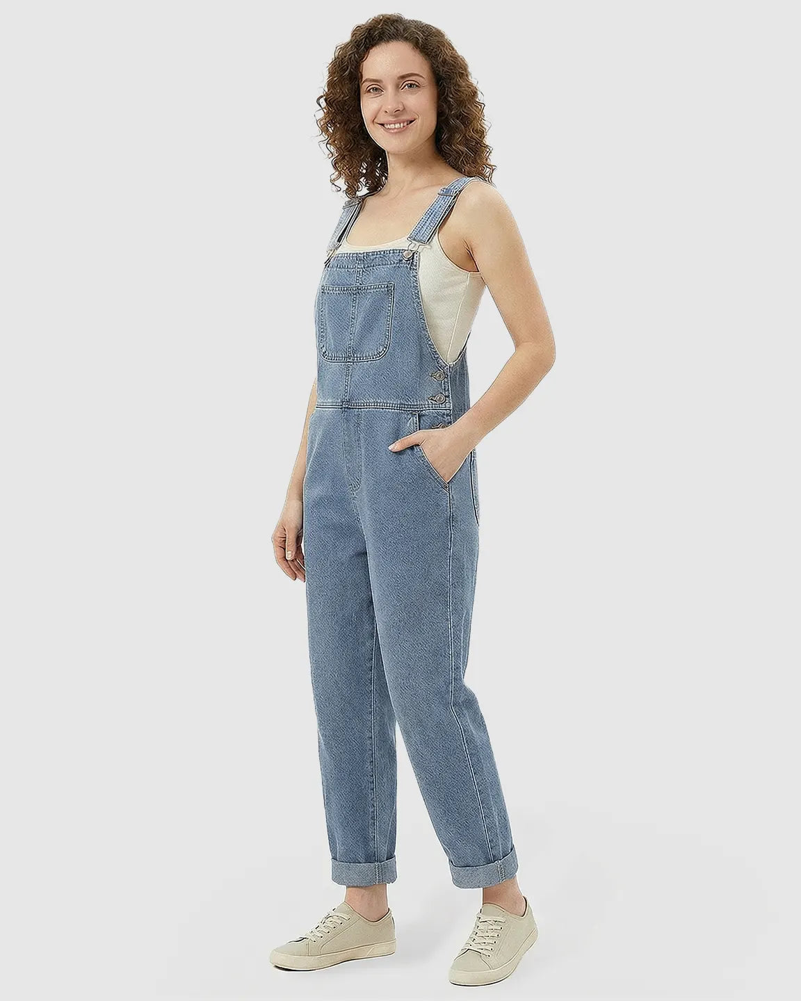 Smiling woman in a light wash denim maternity jumpsuit with adjustable straps, a front pocket, and rolled cuffs.