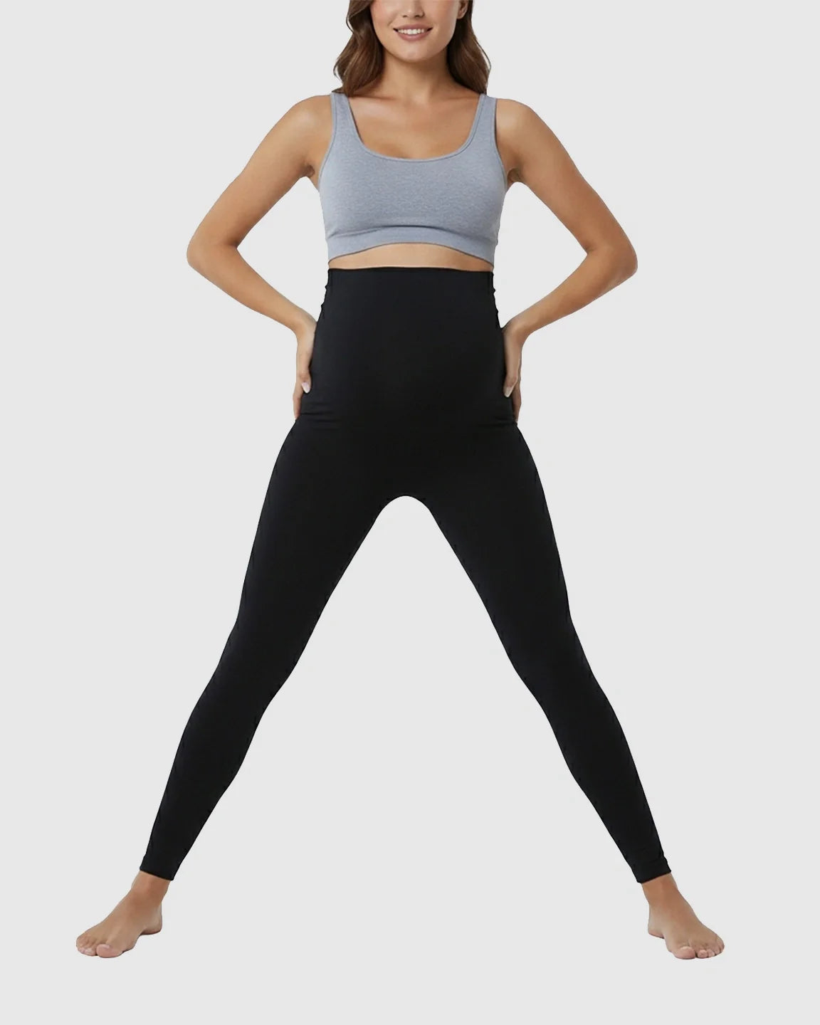 Woman in black high-waist maternity support leggings and a gray sports bra, hands on hips.