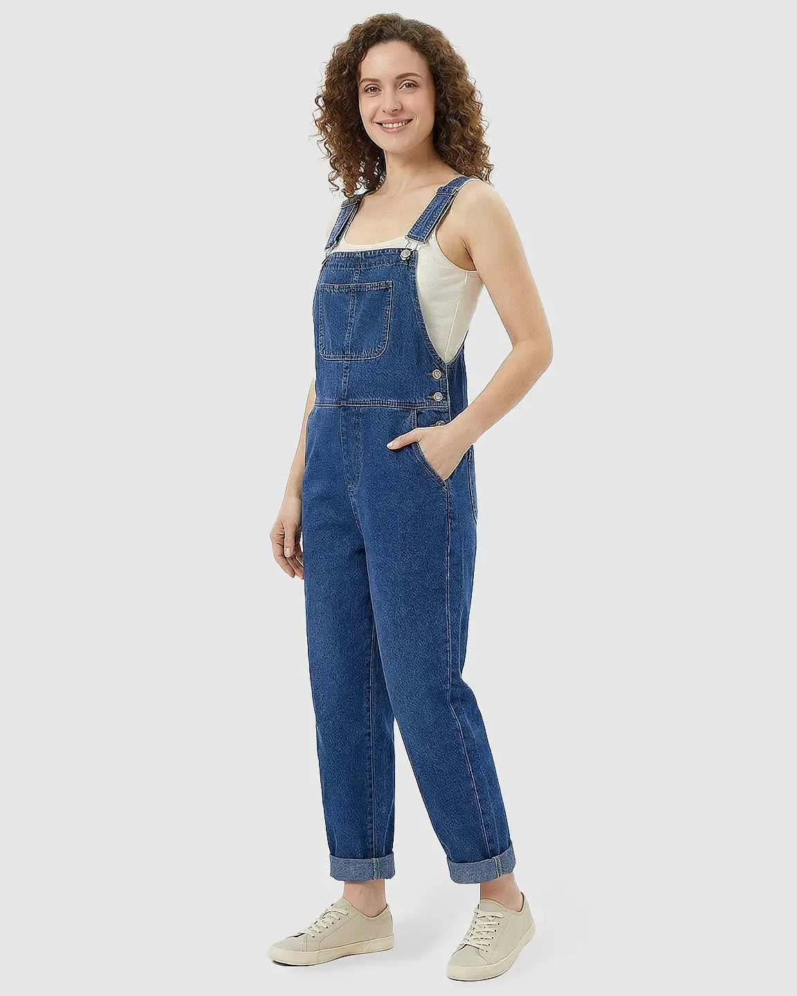 Smiling woman in a loose-fit blue denim maternity jumpsuit with adjustable straps and rolled cuffs.