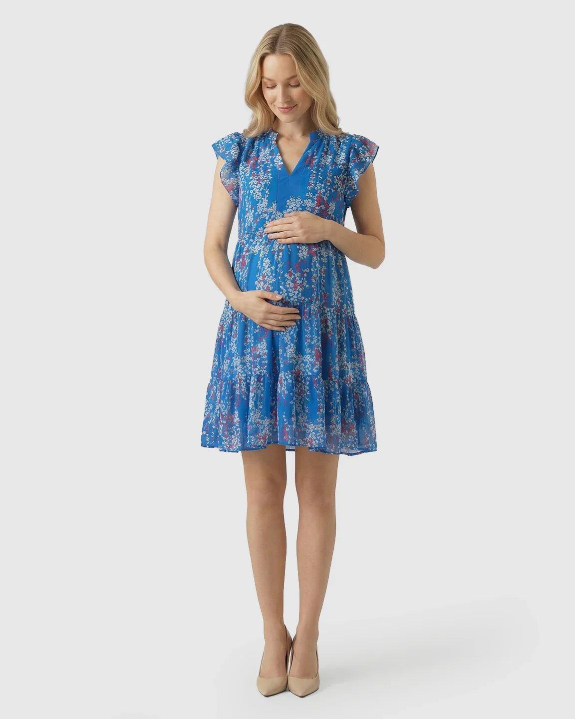 Blonde pregnant woman in a blue floral V-neck A-line maternity dress with flutter sleeves, hands on bump.