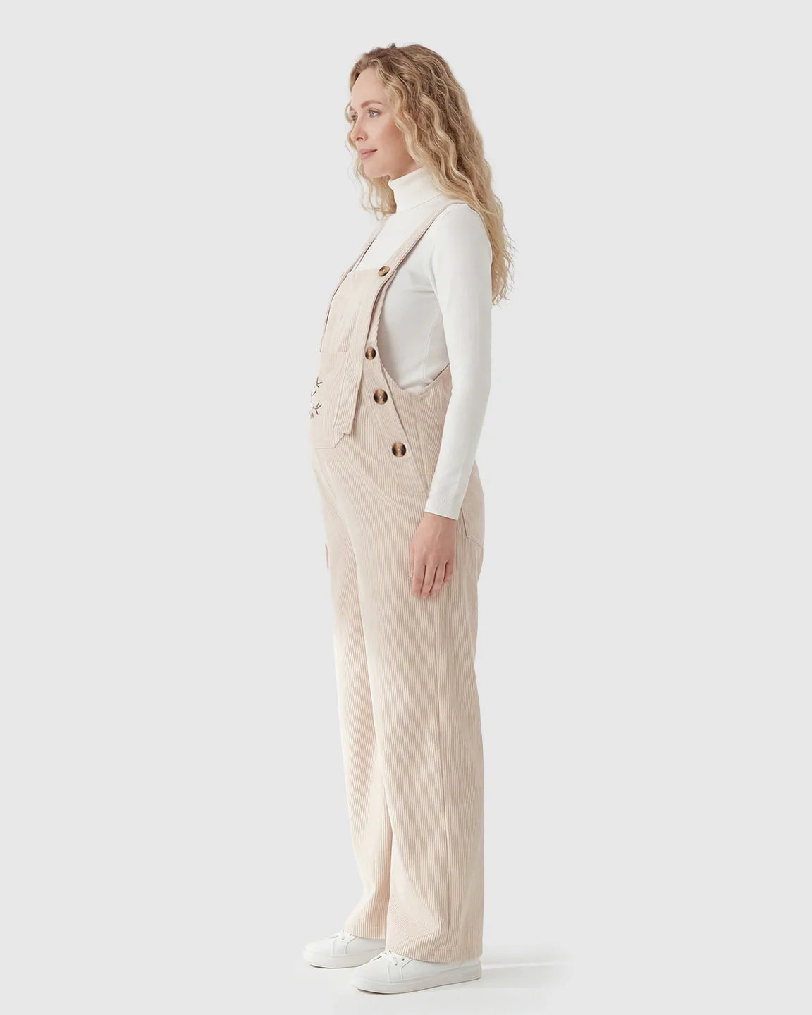 Pregnant woman in side profile wearing beige corduroy maternity overalls with adjustable straps over a white turtleneck.