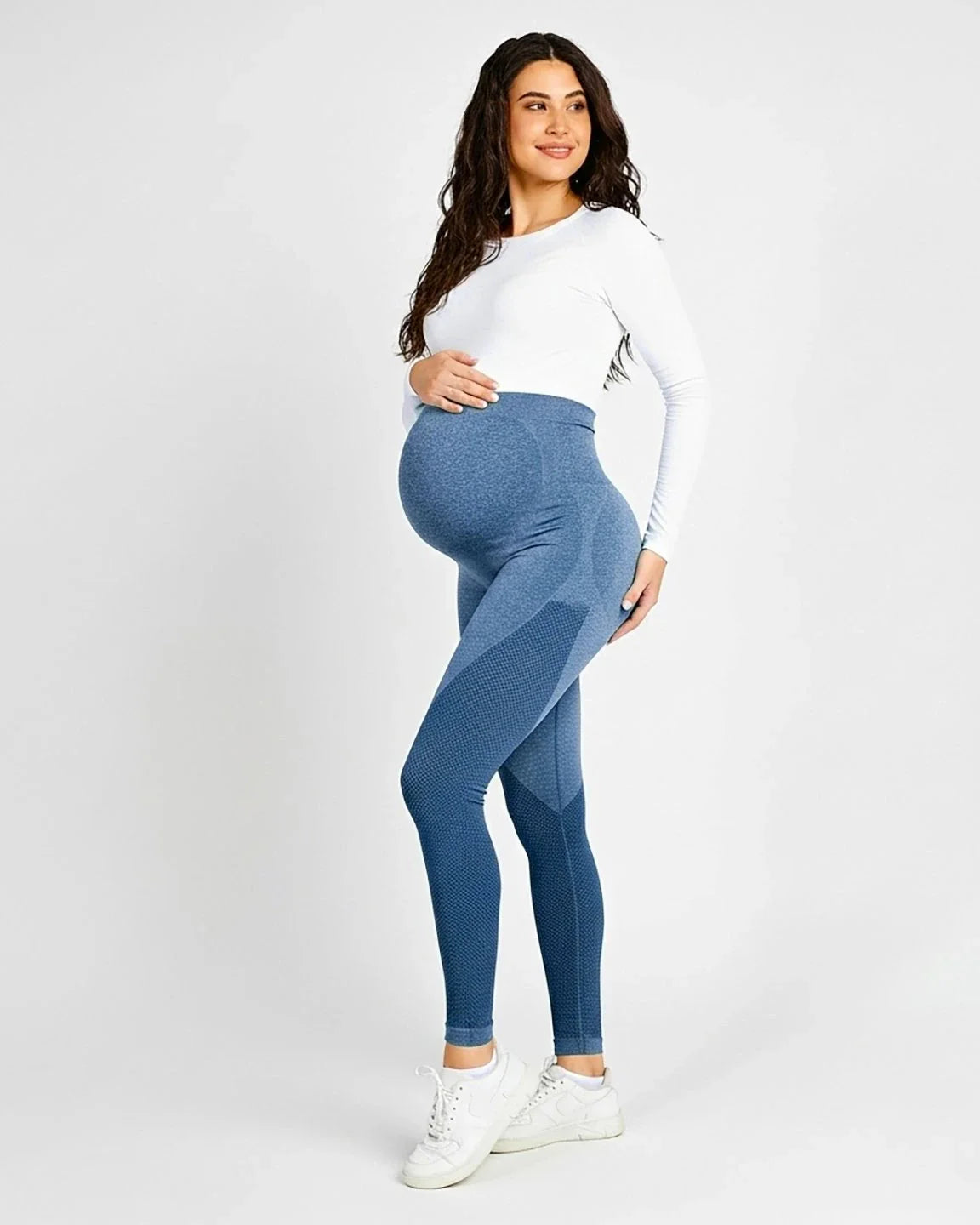 Smiling pregnant woman in blue high-elastic, body-hugging maternity leggings with textured details and a white long-sleeve top.