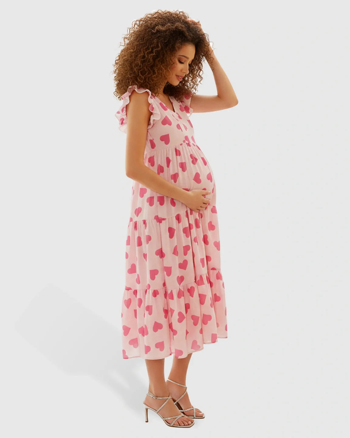 Pregnant woman in a pink chiffon tiered midi dress with red heart print, flutter sleeves, and silver heels.