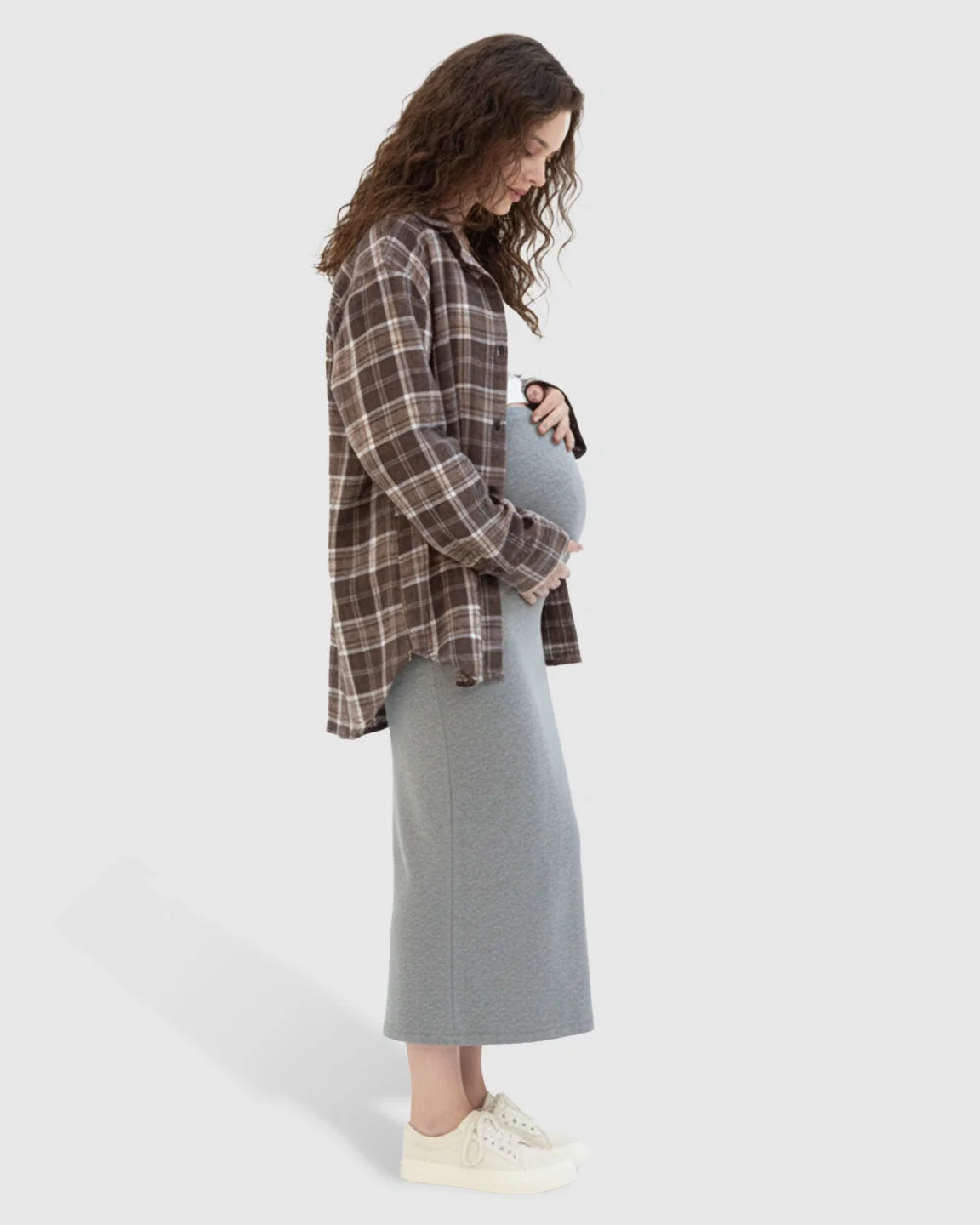 Pregnant woman in a gray midi skirt and brown plaid shirt, gently holding her bump.