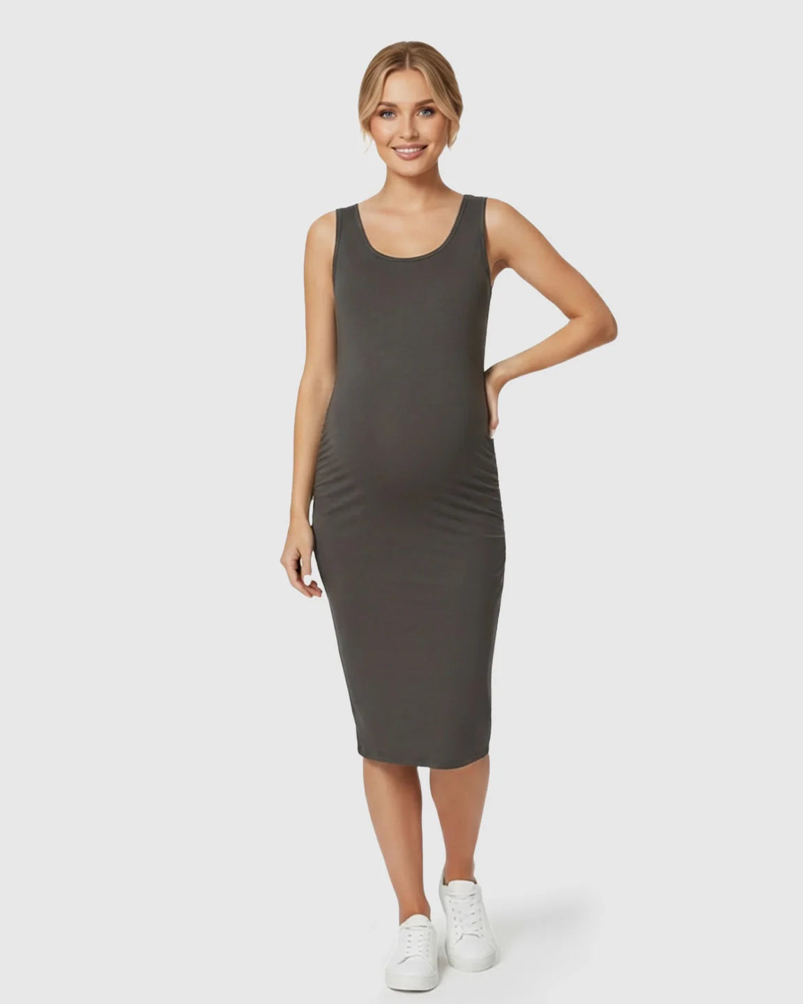 Smiling pregnant woman in a dark gray sleeveless bodycon maternity dress with side pleats and white sneakers.