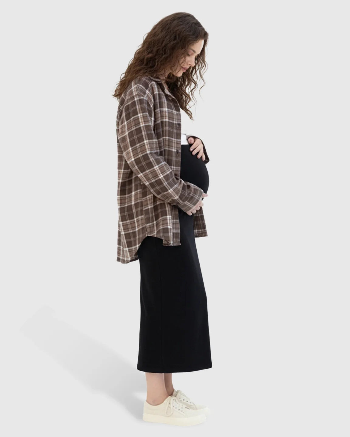 Pregnant woman in a black midi skirt and brown plaid shirt, gently holding her bump.