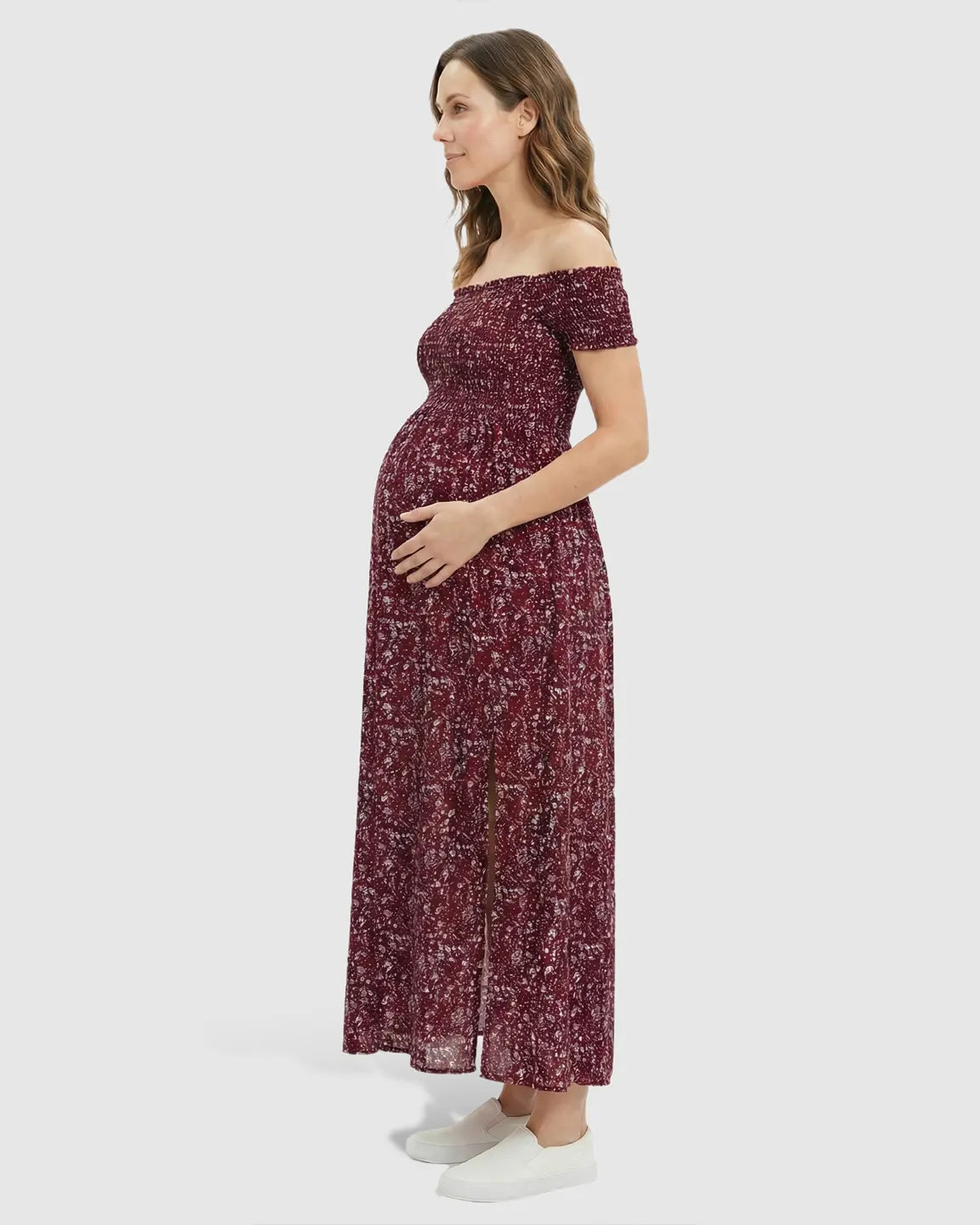 Pregnant woman in a red floral off-shoulder maternity maxi dress with a side slit, holding her bump.