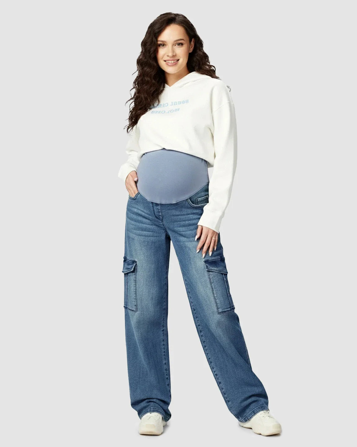 Smiling pregnant woman in blue wide-leg maternity cargo jeans with an over-bump panel and a white hoodie.