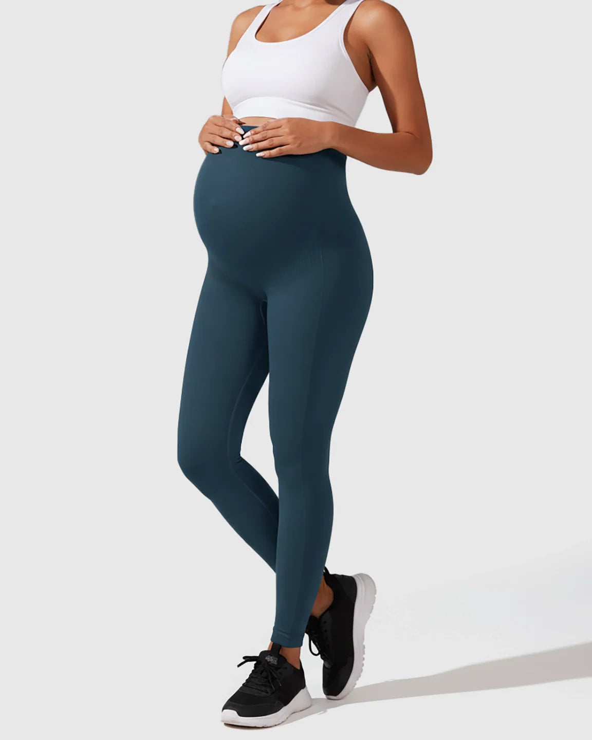 Pregnant woman in seamless, high-waisted teal maternity leggings and a white sports bra, hands on bump.