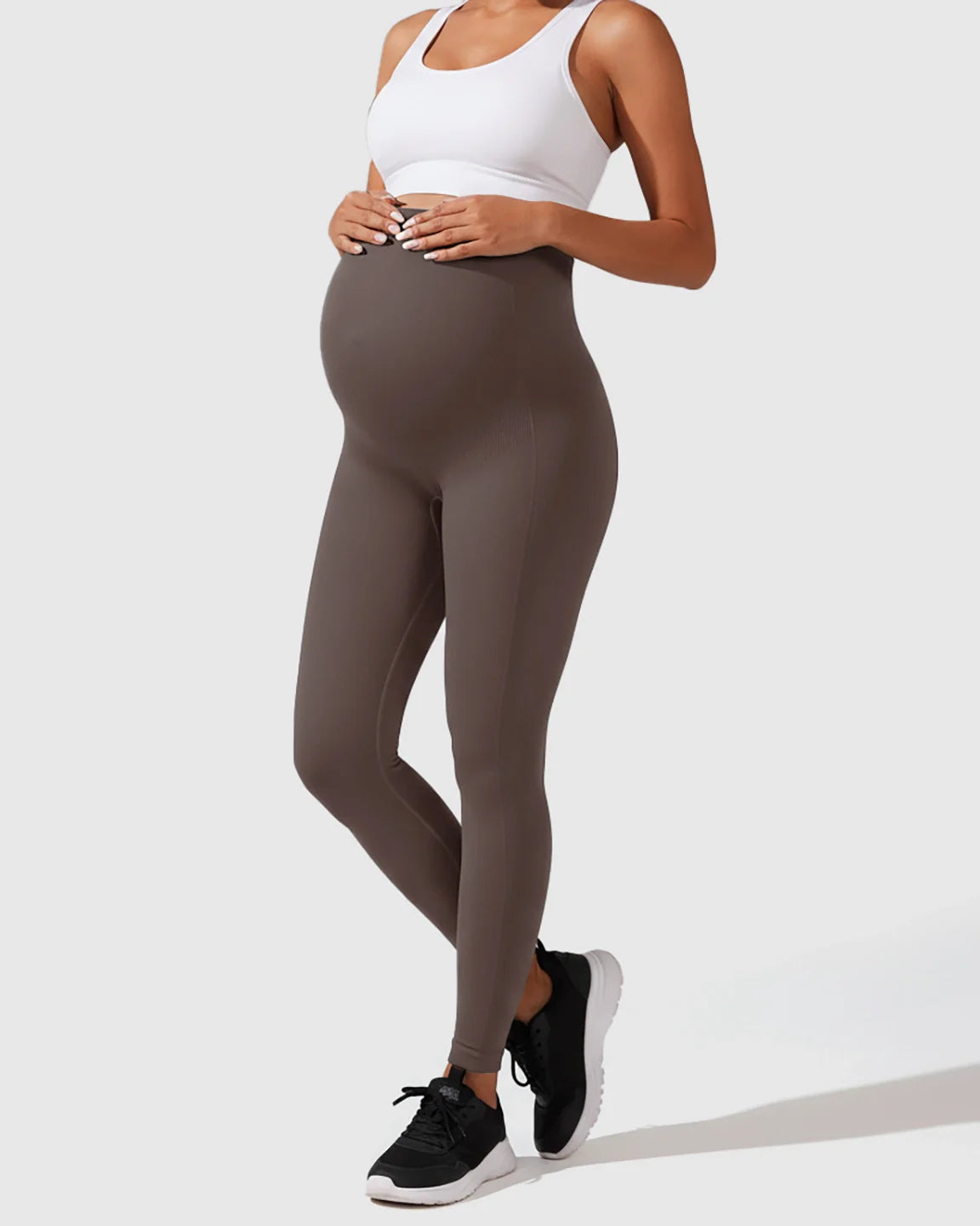 Pregnant woman in seamless, high-waisted taupe maternity leggings and a white sports bra, holding her bump.