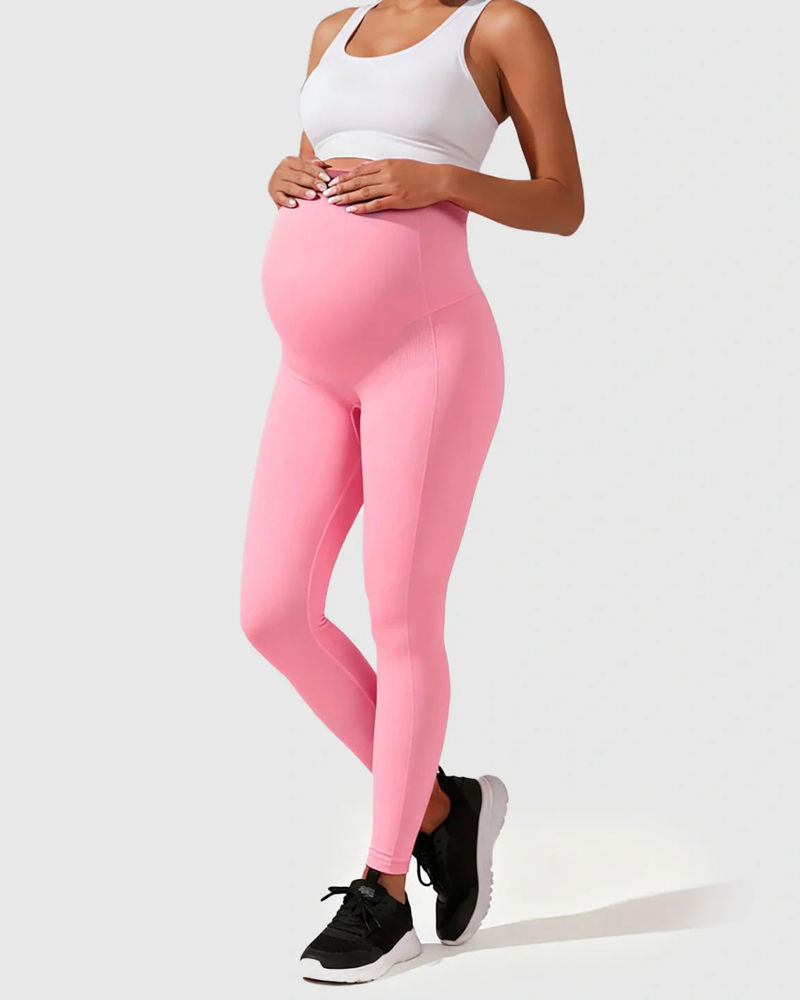 Pregnant woman in pink seamless high-waisted maternity leggings and a white sports bra, cradling her bump.
