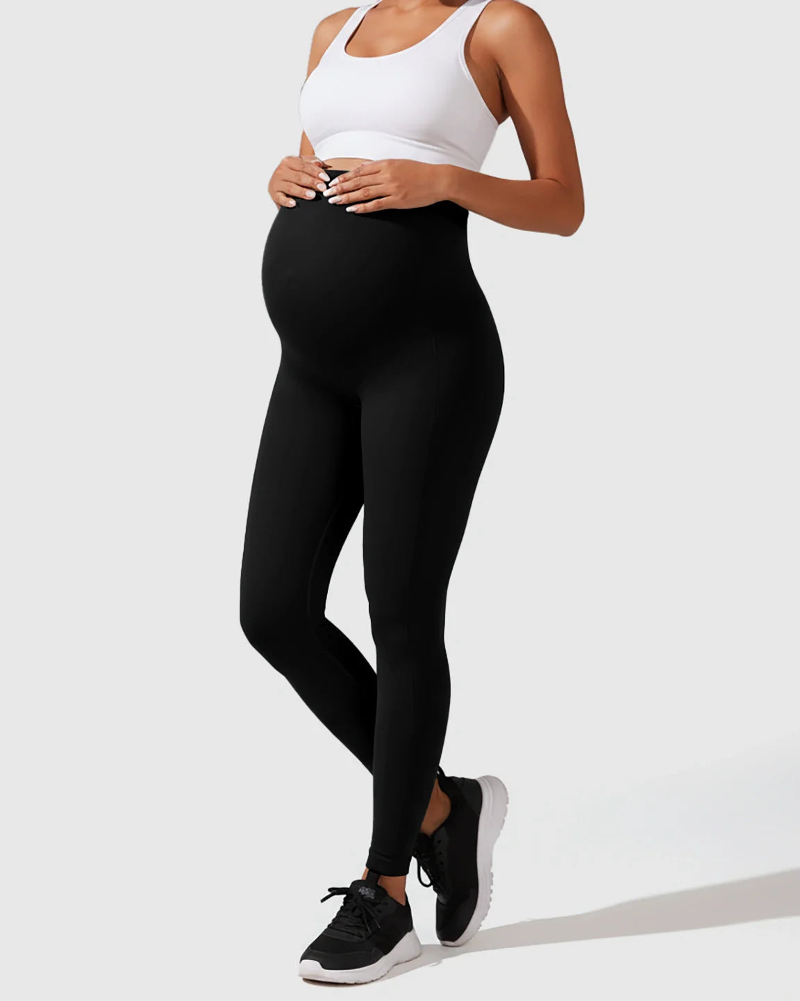 Pregnant woman in black seamless M-shape maternity leggings with full-belly support and a white sports bra.