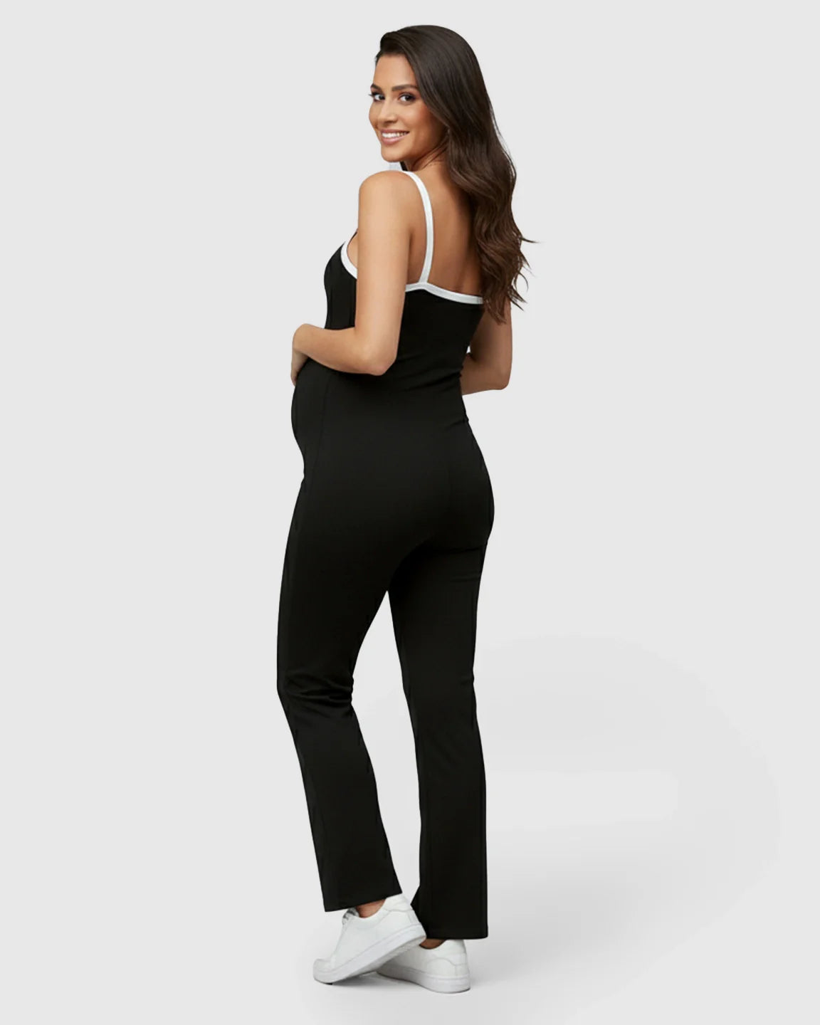 Pregnant woman smiling, wearing a black sleeveless maternity jumpsuit with white trim and white sneakers.