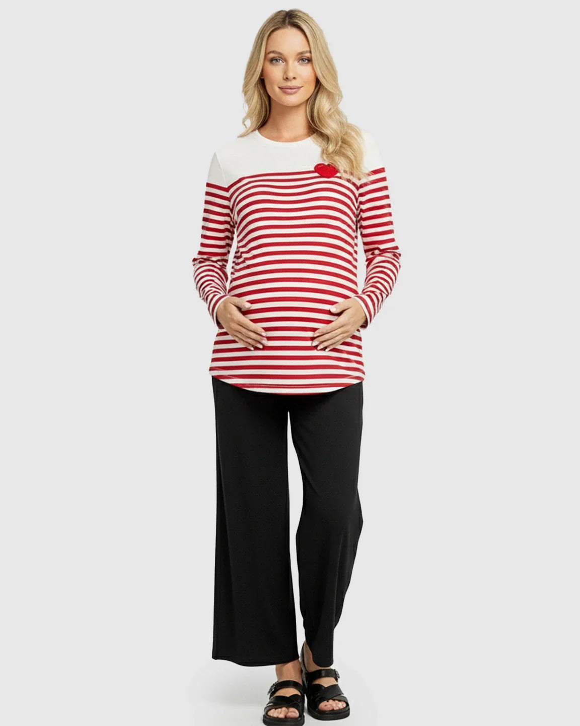 Blonde pregnant woman wearing a red and white striped long-sleeve maternity top with a red heart and black pants.
