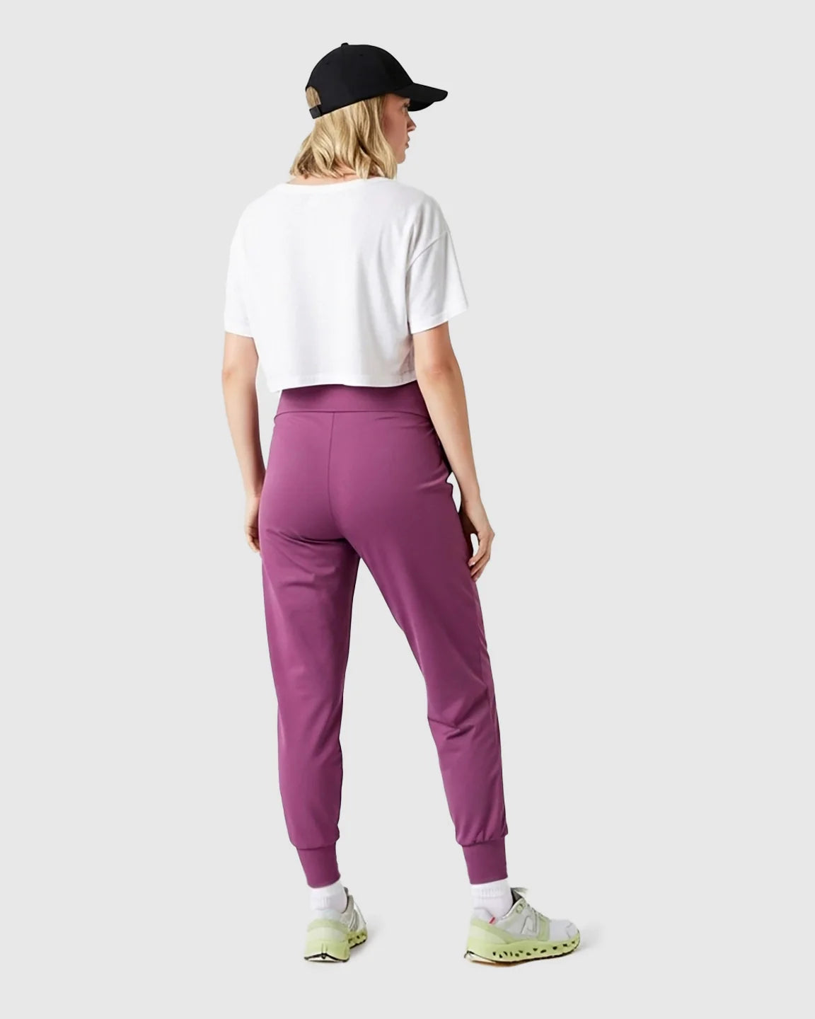 Woman from behind wearing supportive plum maternity joggers, white crop top, black cap, and sneakers.