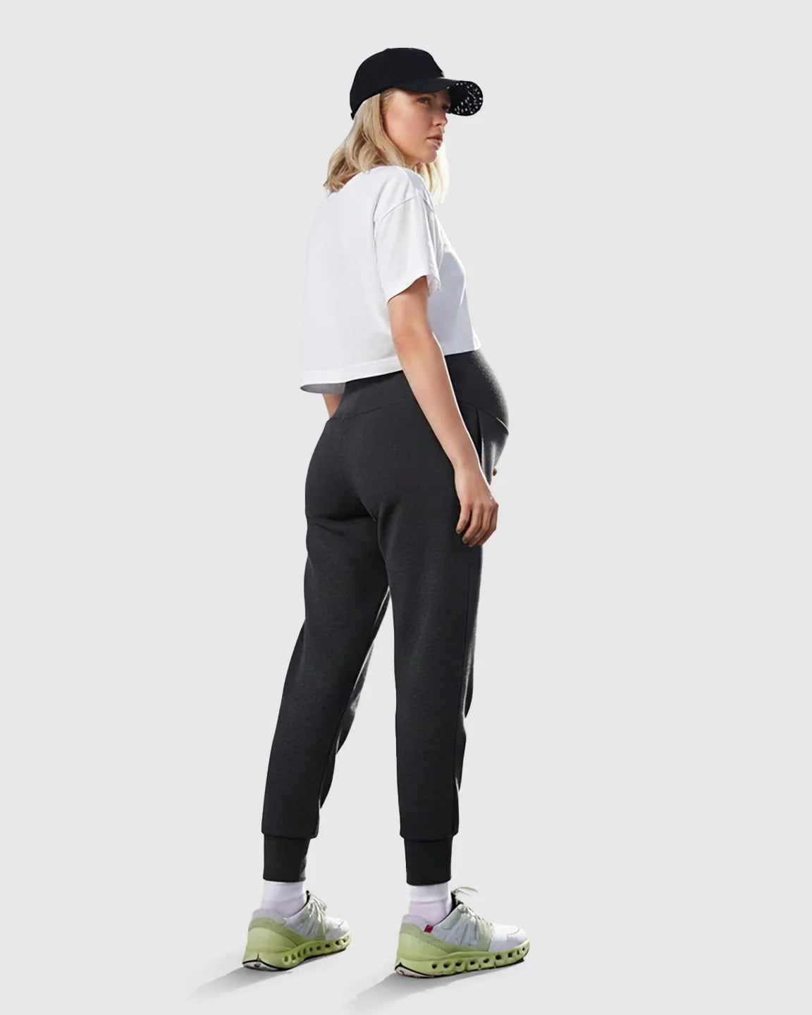 Woman in dark grey supportive slim-fitting maternity joggers, white crop top, and black cap.
