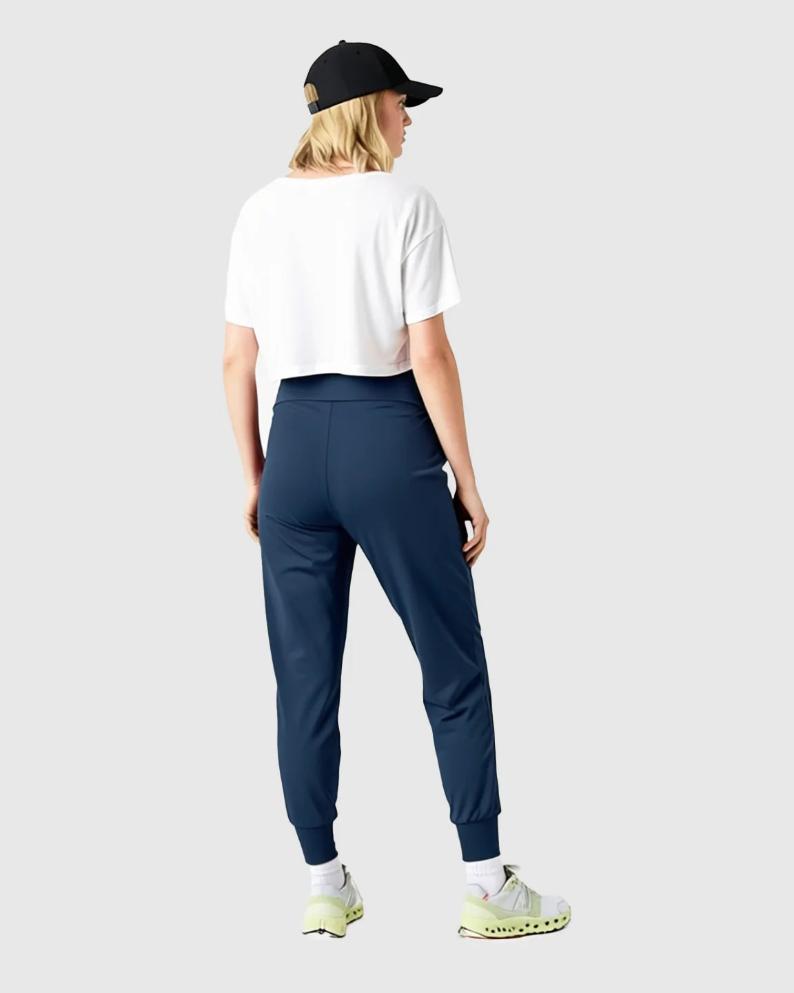 Woman from behind wearing navy supportive slim-fitting maternity joggers, a white crop top, and a black cap.