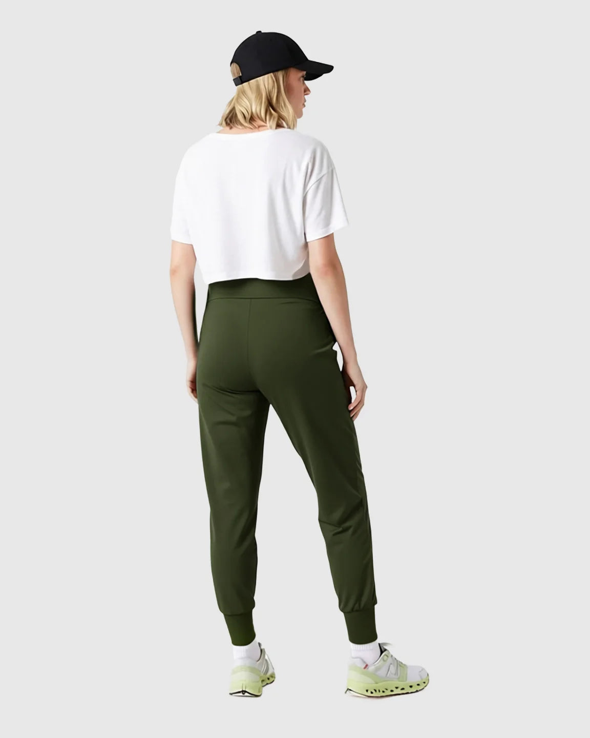 Woman from behind wearing olive green supportive slim-fitting maternity joggers, a white crop top, and a black baseball cap.