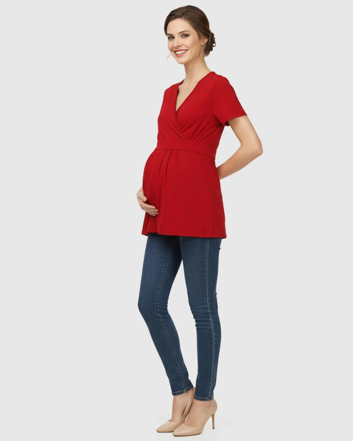 Smiling pregnant woman in a red V-neck wrap-style maternity top with an adjustable waist tie, blue jeans, and heels.