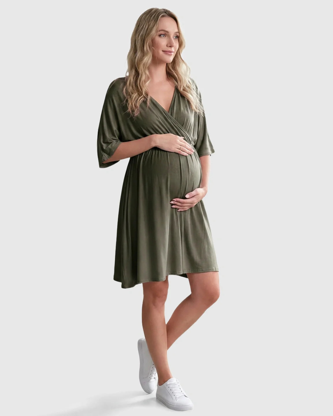 Blonde pregnant woman in a flowy olive green cross-front maternity midi dress with 3/4 sleeves and white sneakers.
