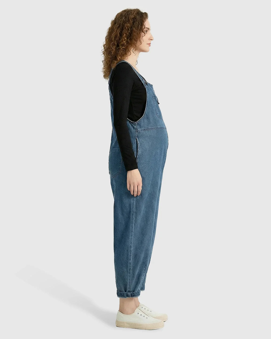 Pregnant woman in loose-fit blue denim maternity overalls with adjustable straps and a black long-sleeve shirt.