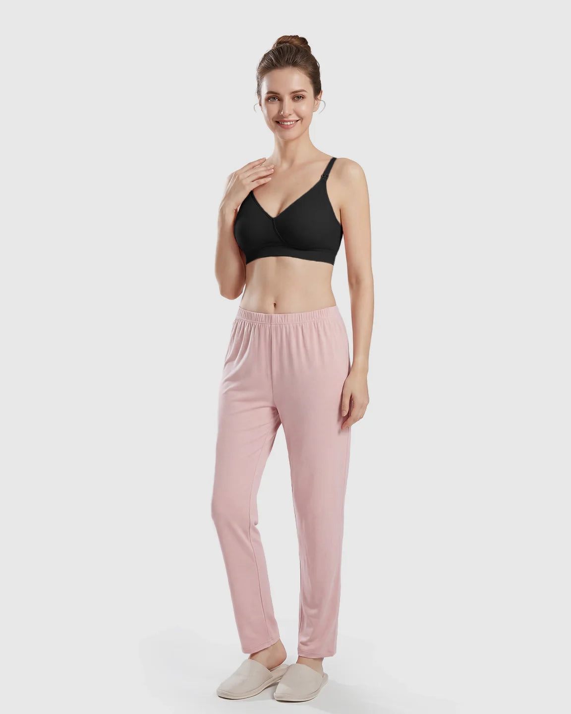 Smiling woman in a black seamless cross-front wireless nursing bra and pink lounge pants.