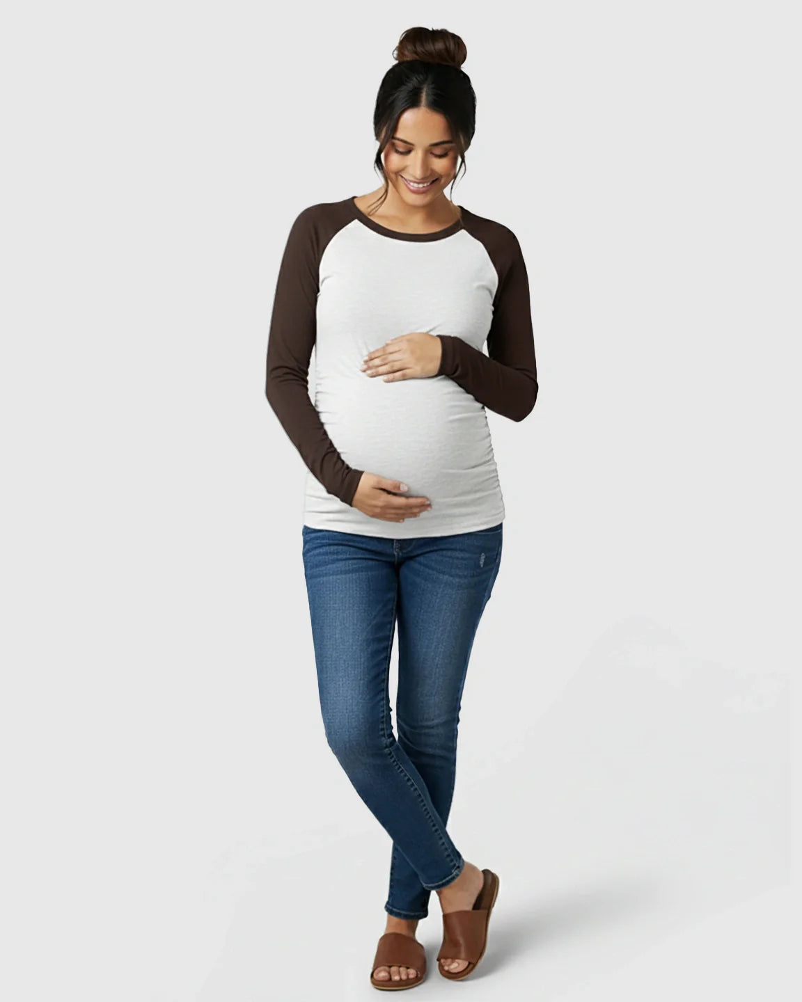 Smiling pregnant woman in a white and brown raglan long-sleeve maternity top with side ruching, blue jeans, and brown sandals.