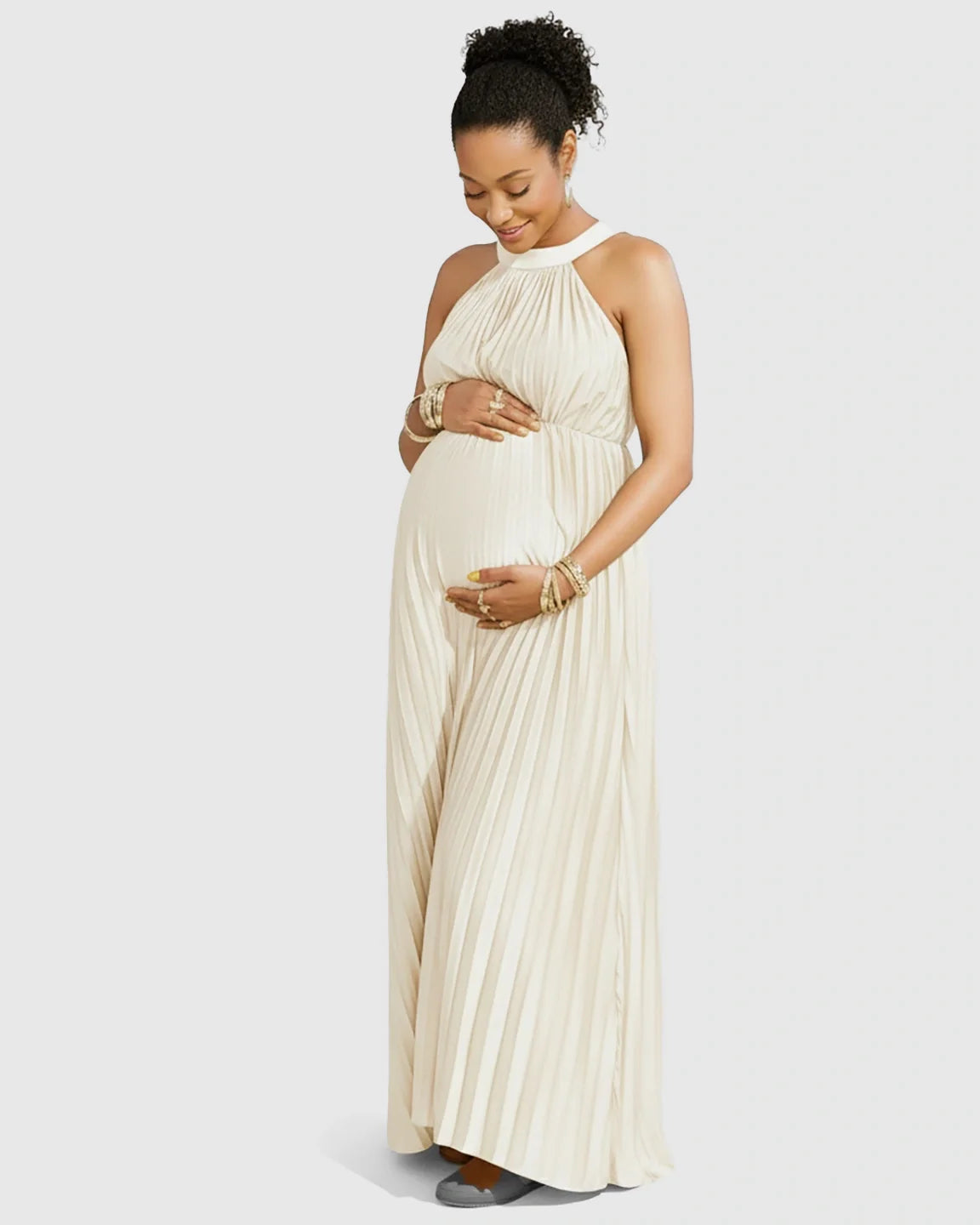 Pregnant woman in an elegant ivory faux silk halter neck pleated maternity maxi dress, hands on belly.