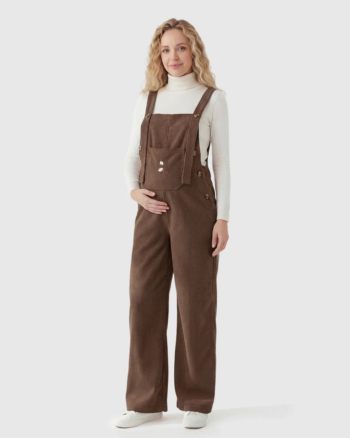 Pregnant woman in brown corduroy maternity overalls with adjustable straps and wide legs over a white turtleneck.
