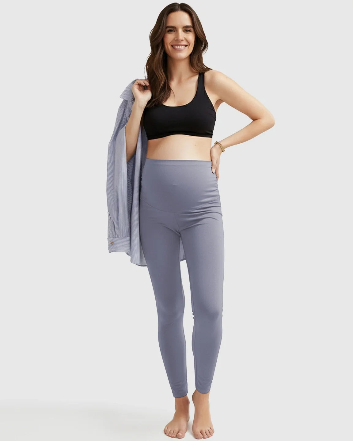 Smiling pregnant woman in ultra-soft grey maternity midi leggings and a black bra, holding a striped shirt.