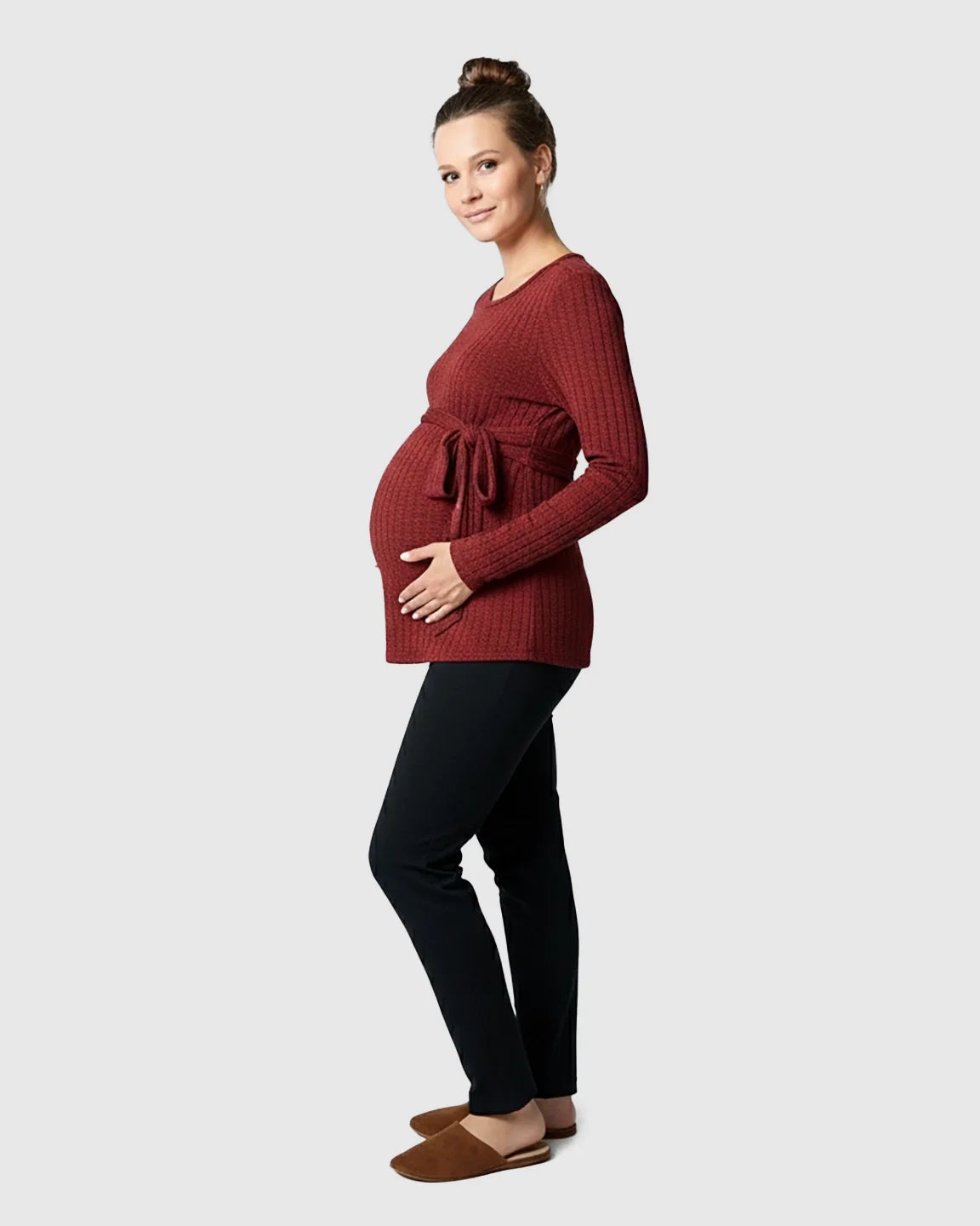 Maternity model in a ribbed, rust-colored tie-waist long sleeve sweater, black pants, and brown slippers.