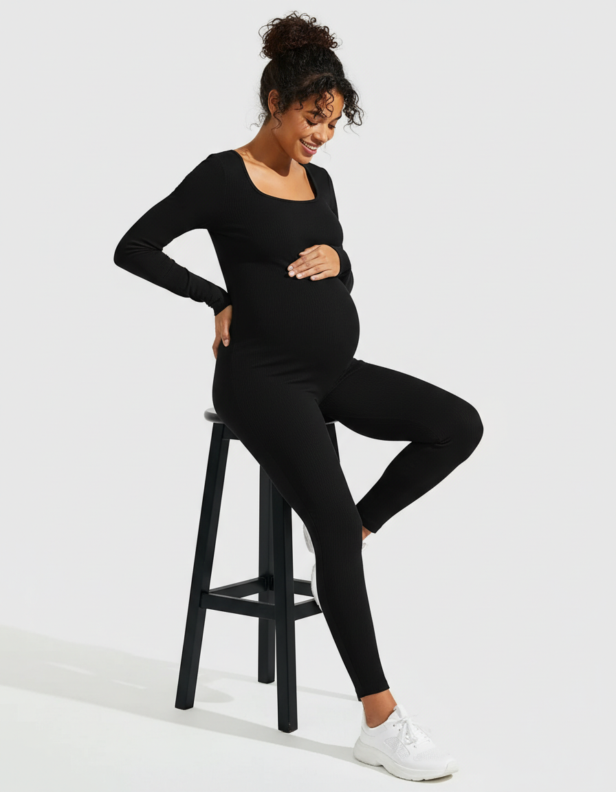 Pregnant woman in a black ribbed long-sleeve maternity jumpsuit with a square neckline, sitting on a stool.
