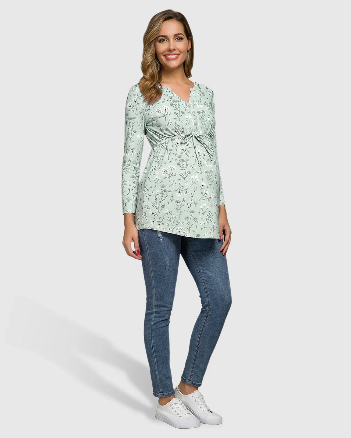 Smiling pregnant woman in a mint green floral long-sleeve maternity tunic with a notch neckline and drawstring waist, paired with jeans.