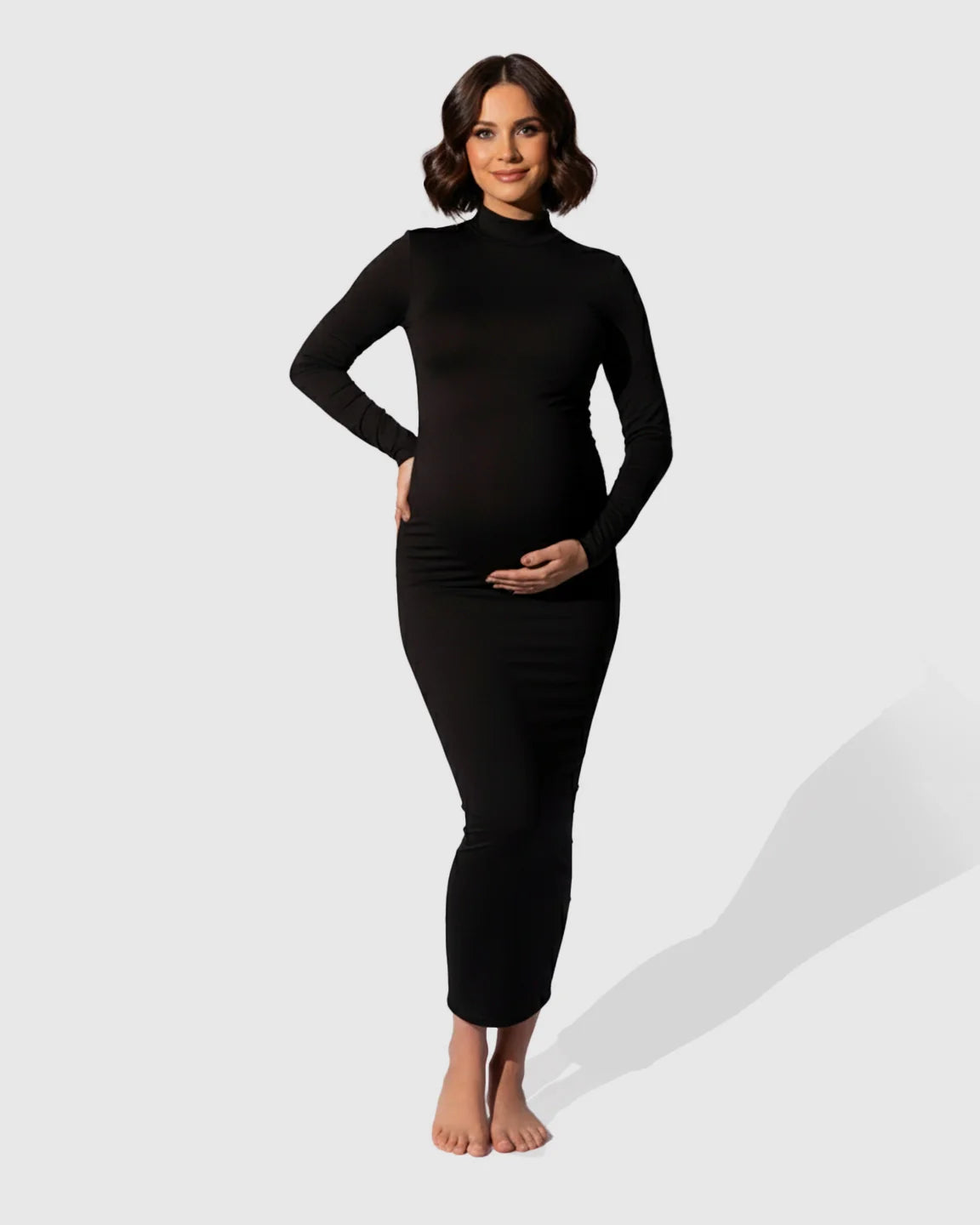 Pregnant woman in a black mock-neck, long-sleeve bodycon maternity maxi dress, gently cradling her bump.