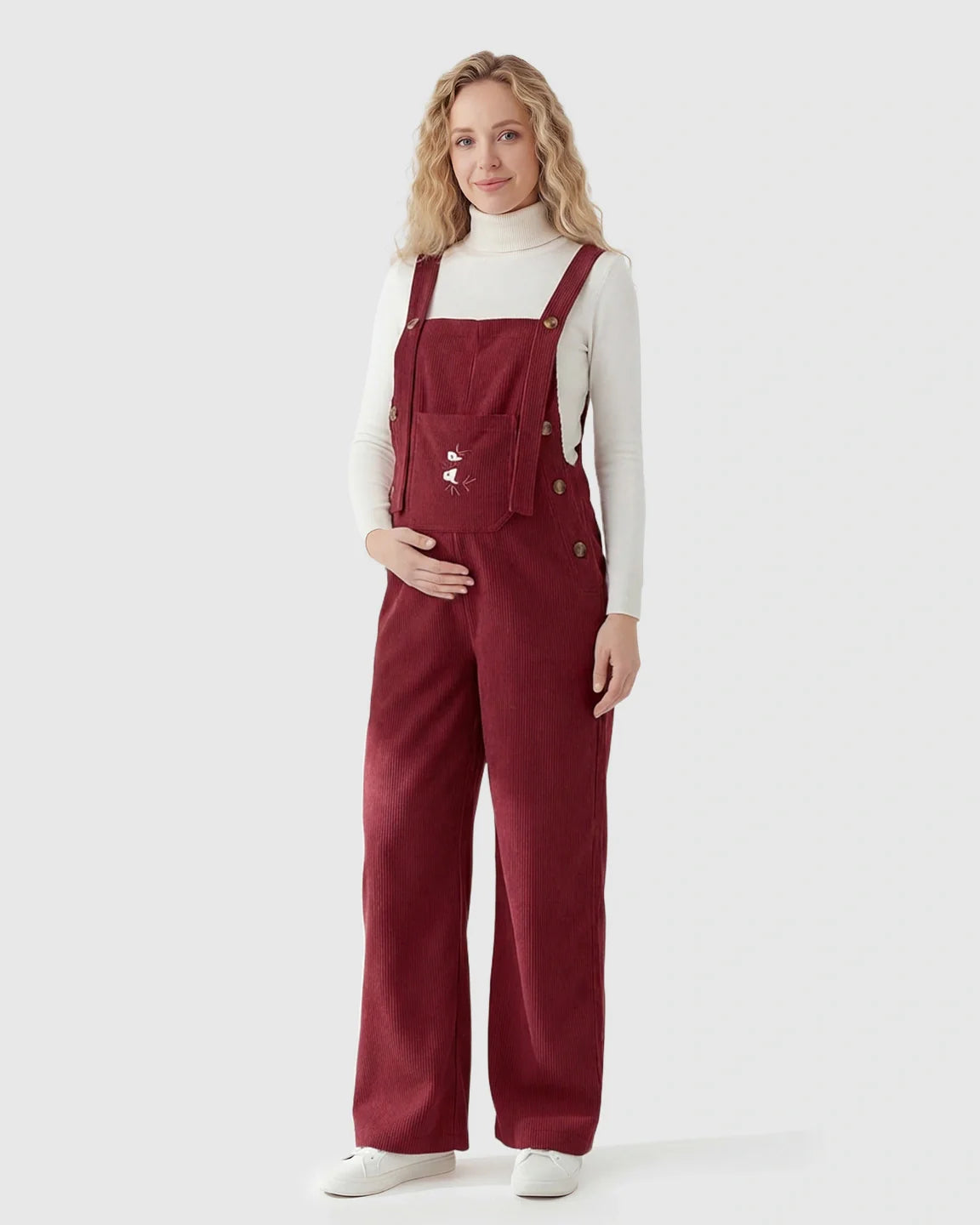 Pregnant woman in red corduroy maternity overalls with adjustable straps and white turtleneck.